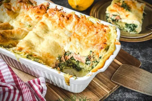 Herzhafte Lachs Spinat Lasagne, goldbraun überbacken, dampft in einer weißen Auflaufform. Ein Stück auf einem Teller zeigt die Schichten.