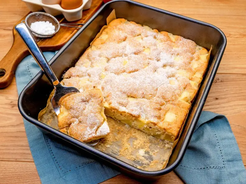 Ein Stück vom schneller saftiger apfelkuchen vom blech wird mit einem Kuchenheber herausgehoben. Mit Puderzucker bestäubt.